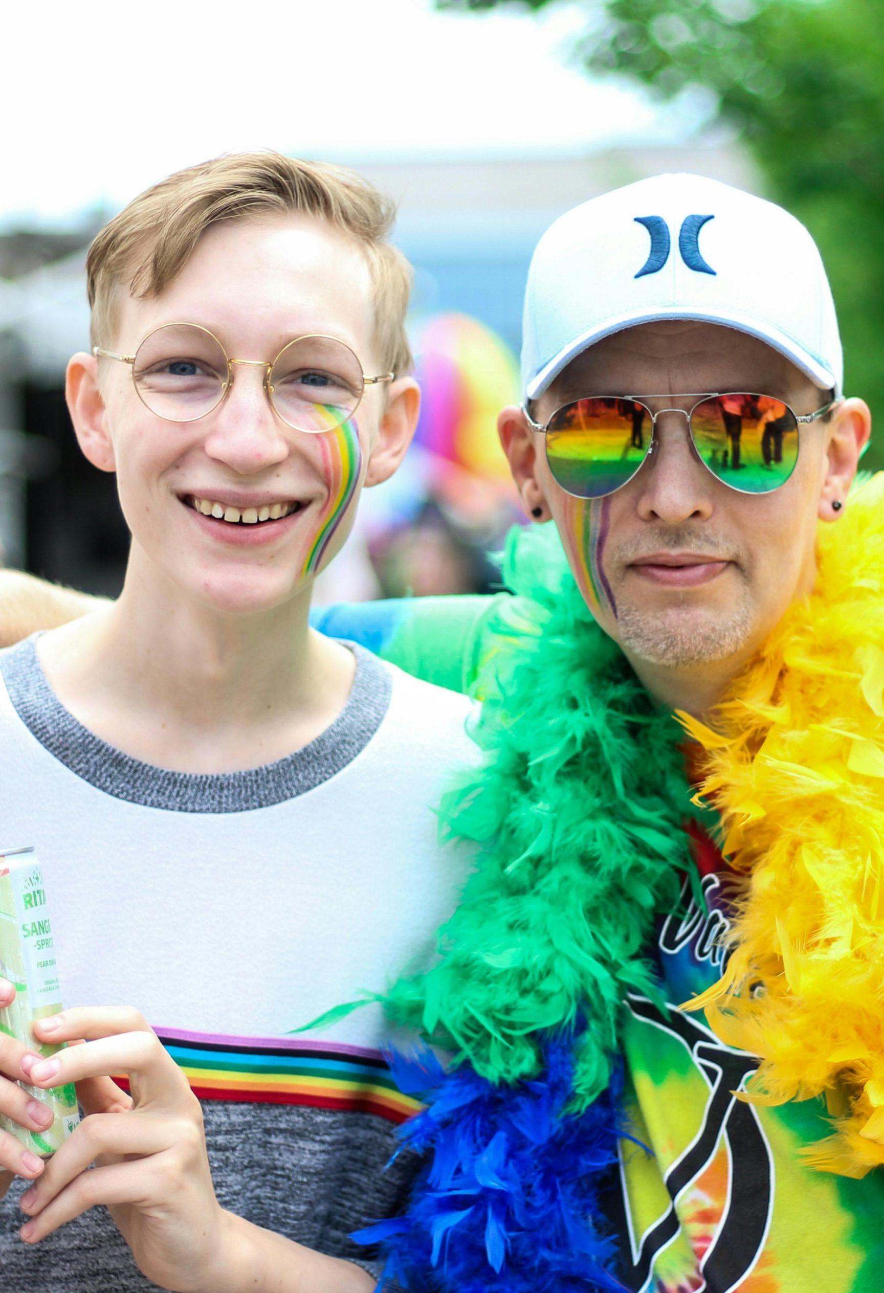 A man and a teenage boy pose for a photo. They both have rainbows painted on their cheeks, and the man is wearing a rainbow feather boa.